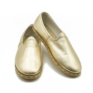 May include: A pair of gold leather slip-on shoes. The shoes have a rounded toe and a stitched sole. The leather has a metallic sheen. The shoes are displayed on a white background.
