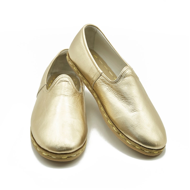 May include: A pair of gold leather slip-on shoes. The shoes have a rounded toe and a stitched sole. The leather has a metallic sheen. The shoes are displayed on a white background.