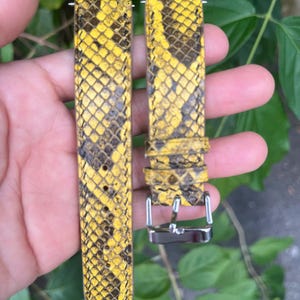May include: Yellow and black snakeskin-patterned watch straps with silver-tone hardware. The straps are made of a textured material and feature a repeating scale design. The watch straps are shown against a green leafy background.