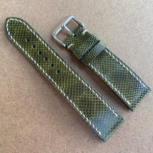 以下が含まれることがあります： 白いステッチが付いた緑色の革製の腕時計のストラップ。ストラップはテクスチャ加工された革製で、銀色のバックルが付いています。