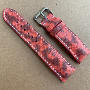 Puede incluir: Una correa de reloj de cuero con estampado rojo y marrón con costuras blancas. La correa tiene una hebilla plateada.