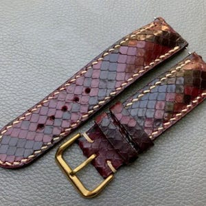 Puede incluir: Una correa de reloj de piel de serpiente marrón y negra con herrajes dorados. La correa tiene un borde cosido y un patrón texturizado.