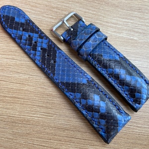 May include: Two blue and black patterned leather watch straps with blue stitching. The straps are laid out flat on a wooden surface.