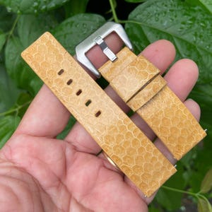 Pode incluir: Uma pulseira de relógio de couro marrom claro com um padrão de favo de mel. A pulseira tem uma fivela prateada e é mostrada em uma mão.