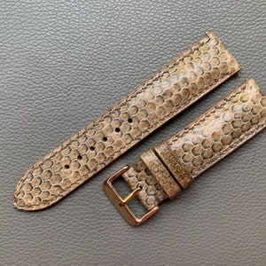 Puede incluir: Una correa de reloj de cuero marrón con un patrón texturizado. La correa tiene una hebilla dorada.