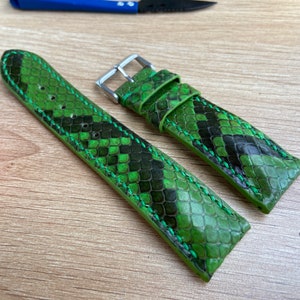 Puede incluir: Dos correas de reloj de piel de serpiente verde con patrones negros contrastantes y costuras verdes. Las correas están dispuestas sobre una superficie de madera.