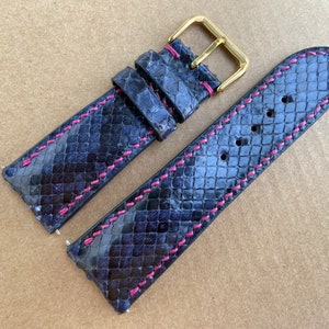 Peut inclure: Un bracelet de montre bleu foncé en peau de serpent avec des coutures roses. Le bracelet a une boucle dorée.