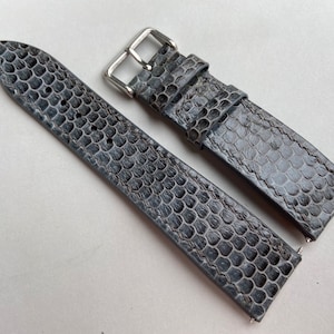 Puede incluir: Una correa de reloj de cuero gris con un patrón texturizado de serpiente. La correa tiene una hebilla plateada.