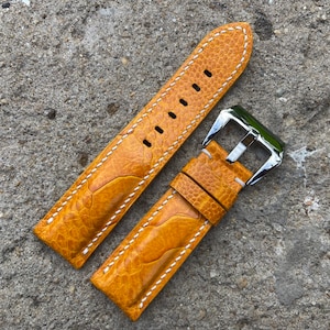 Puede incluir: Una correa de reloj de cuero amarillo con costuras blancas y una hebilla plateada.