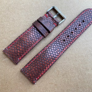 May include: A brown leather watch strap with red stitching. The strap has a textured surface and a silver buckle.