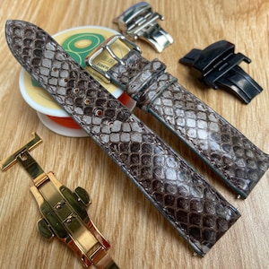 Może przedstawiać: Paski do zegarków ze wzorem skóry węża w odcieniach brązu i szarości. Na zdjęciu widoczne są również zapięcia do zegarków w kolorze złotym, srebrnym i czarnym oraz szpulka nici. Paski te to luksusowy przedmiot.