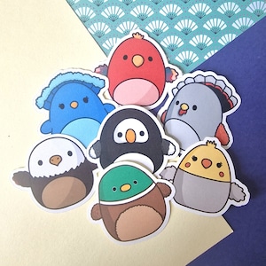 Può includere: Una collezione di adesivi di uccelli colorati con illustrazioni in stile cartone animato. Gli adesivi presentano diversi uccelli, tra cui un uccello rosso, un uccello blu, un pinguino e un'anatra. Gli adesivi sono disposti su uno sfondo giallo chiaro e blu.