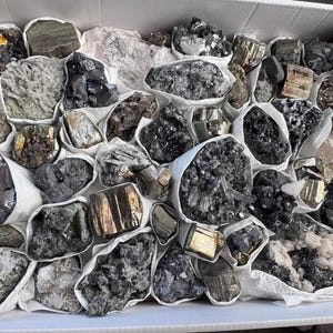May include: A collection of various grey, black, and gold crystals in individual white paper bags. The crystals are arranged in a box.