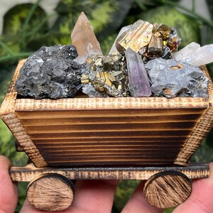 May include: A wooden cart filled with various crystals and minerals. The cart is made of light brown wood with dark brown accents. The crystals include quartz, pyrite, and amethyst. The cart has two wooden wheels.