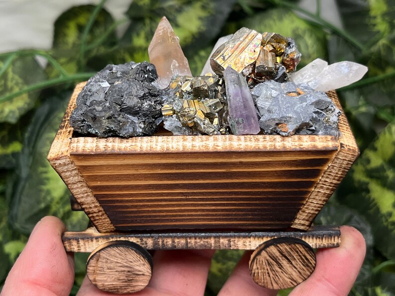 May include: A wooden cart filled with various crystals and minerals. The cart is made of light brown wood with dark brown accents. The crystals include quartz, pyrite, and amethyst. The cart has two wooden wheels.