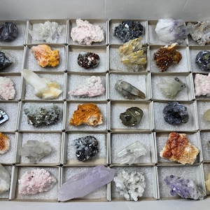 Könnte beinhalten: Eine Sammlung verschiedener farbenfroher Mineralproben, die in einzelnen Pappschachteln präsentiert werden. Die Mineralien weisen eine Reihe von Farben auf, darunter Rosa, Orange, Lila und Klar, mit unterschiedlichen Texturen und Kristallformationen. Die Schachteln sind in einem größeren Pappbehälter angeordnet.