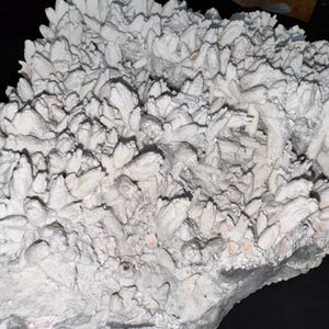 Op de afbeelding: Een grote, gebroken witte cluster van kristalformaties. De kristallen hebben een getextureerd, gelaagd uiterlijk, met veel kleine, puntige uiteinden. De algemene vorm is onregelmatig en organisch, met een ruw, ongelijkmatig oppervlak. De kristallen zijn dicht opeengepakt.