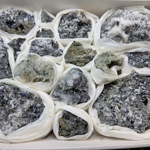 May include: A collection of various mineral specimens, including gray, silver, and green hues, are displayed in white paper packaging within a cardboard box. The minerals have a crystalline structure.