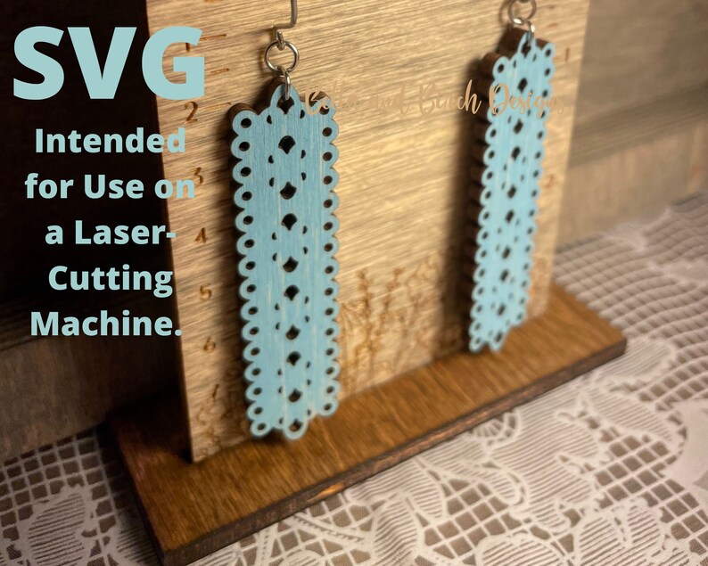May include: A pair of light blue laser cut earrings with a delicate lace pattern. The earrings are displayed on a wooden stand with a ruler. SVG Intended for Use on a Laser-Cutting Machine.