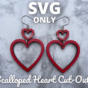 Puede incluir: Un par de pendientes de corazón rojo festoneado con un corazón más pequeño recortado dentro de cada corazón más grande. Los pendientes están hechos de madera y tienen ganchos plateados.