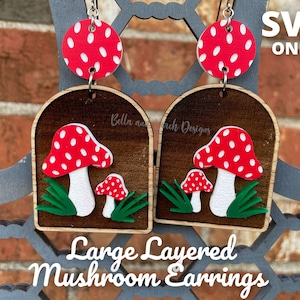 May include: Large layered mushroom earrings with red and white polka dot tops. The earrings feature a wooden arch design with a red and white mushroom and green grass. The text "Large Layered Mushroom Earrings" is displayed.
