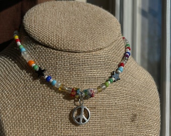 Puede incluir: Un collar de cuentas de colores con un dije de signo de paz. El collar presenta cuentas blancas, azules, verdes, amarillas, rojas y negras, así como dijes de estrellas plateadas.