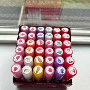 Könnte beinhalten: Eine Auswahl an Lipgloss-Tuben in verschiedenen Farben, jede mit einer anderen Fruchtillustration auf der Oberseite. Früchte wie Erdbeeren, Pfirsiche, Kirschen und Blaubeeren sind abgebildet. Die Tuben sind in einer schwarzen Box angeordnet.