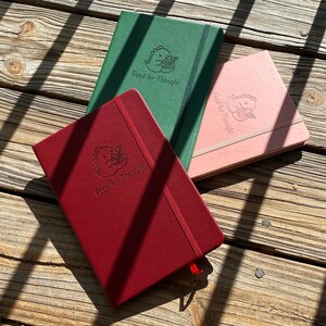 Op de afbeelding: Drie hardcover notitieboekjes met een rode, roze en groene omslag. Elk notitieboekje heeft een dinosaurus afbeelding en de tekst "Food for Thought".