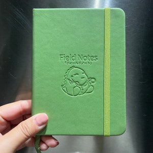 Op de afbeelding: Een groen Field Notes notitieboekje met een groene elastieken sluiting en een reliëf van een dinosaurus die een potlood vasthoudt. De tekst "Field Notes" en "フィールドノート" is reliëf op de voorzijde.