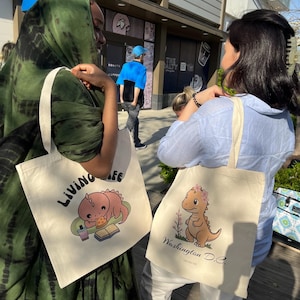 Puede incluir: Dos bolsas de tela con diseños de dinosaurios de dibujos animados. Una bolsa es verde y blanca con el texto "Living Life" y un dinosaurio leyendo un libro. La otra bolsa es beige con un dinosaurio con flores en la cabeza y el texto "Washington D.C."