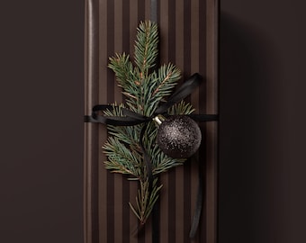 May include: A rectangular gift box wrapped in dark brown paper with vertical black stripes. The package is tied with a thin black ribbon, featuring a green pine sprig and a small, round, dark brown glittery ornament attached to the center.