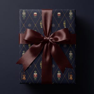 May include: A rectangular gift box wrapped in dark navy blue paper featuring a repeating gold diamond grid pattern with illustrations of nutcrackers, drums, trumpets, violins, and starbursts, finished with a wide, dark brown satin ribbon bow.