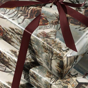 May include: A stack of three rectangular gift boxes wrapped in paper featuring a vintage winter scene with a horse-drawn sleigh and snowy houses. The boxes are tied with a dark burgundy satin ribbon and include a matching gift tag.