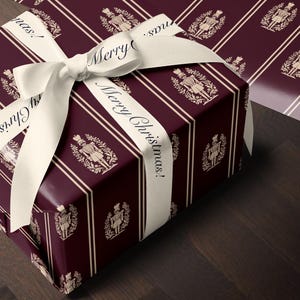 May include: A rectangular gift box wrapped in deep burgundy paper featuring a repeating pattern of cream-colored nutcracker figures within laurel wreaths and vertical stripes, tied with a cream ribbon printed with the text Merry Christmas.