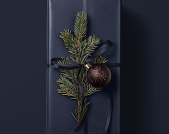 May include: A rectangular gift box wrapped in matte navy blue paper, tied with a thin navy blue satin ribbon. A green pine sprig is tucked under the ribbon, accompanied by a single round, dark brown glittery ornament hanging from the center.