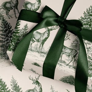 Op de afbeelding: Een rechthoekige geschenkdoos verpakt in crèmekleurig papier met een donkergroen gedrukt patroon van herten en dennenbomen. Het pakket is gestrikt met een breed, donkergroen satijnen lint dat bovenop tot een strik is geknoopt.