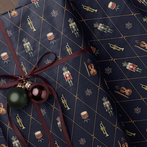 May include: A dark blue gift wrap paper featuring a repeating pattern of nutcrackers, violins, trumpets, drums, and starbursts within a gold diamond grid. The package is tied with a dark red ribbon and two round ornaments in matte green and glossy dark red.