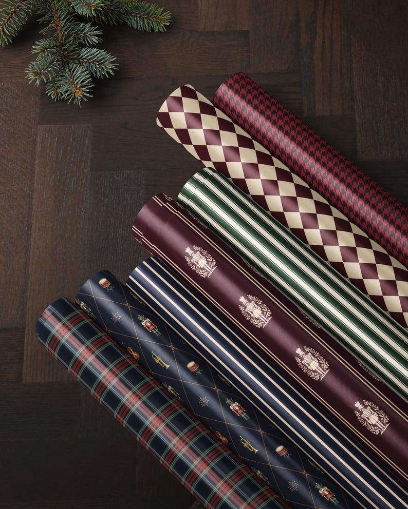 May include: Six rolls of wrapping paper featuring various patterns including a red and cream diamond print, a red houndstooth, green and white stripes, a deep red with gold nutcracker motifs, navy blue stripes, and a navy blue plaid pattern.