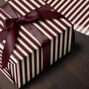 Könnte beinhalten: Eine rechteckige Geschenkbox, eingewickelt in Papier mit einem Muster aus vertikalen dunkelburgunderroten und cremefarbenen Streifen. Die Box ist mit einem breiten, glatten dunkelburgunderroten Satinband umwickelt, das oben zu einer Schleife gebunden ist.