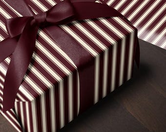 May include: A rectangular gift box wrapped in paper featuring a pattern of vertical dark burgundy and cream-coloured stripes. The box is tied with a wide, smooth dark burgundy satin ribbon that is knotted into a bow on the top surface.