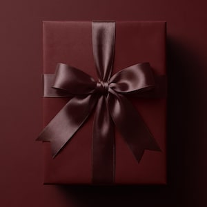 Könnte beinhalten: Eine rechteckige Geschenkbox, eingewickelt in einfarbiges, tiefburgunderrotes Papier. Die Box ist mit einem passenden dunkelburgunderroten Satinband umwickelt, das eine große, mittige Schleife mit zwei langen, spitz zulaufenden Enden bildet.