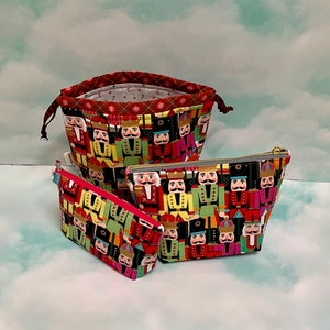 May include: Three fabric pouches with a repeating nutcracker soldier pattern. The largest is a drawstring bag with a red plaid interior. The other two are zippered cosmetic bags. The bags have a red, green, yellow, and black color scheme.