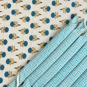 Puede incluir: Un mantel blanco con un patrón repetido de diseños florales azules y una pila de servilletas plegadas turquesas y blancas con estampado de chevrón. Las servilletas están dispuestas diagonalmente sobre el mantel.