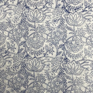 Op de afbeelding: Een witte stof met een blauw bloemenpatroon. Het patroon bevat verschillende bloemen en bladeren, allemaal in een delicate, ingewikkelde stijl.