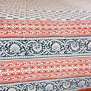 Puede incluir: Un textil con un patrón repetitivo de diseños rojos, azul marino y blancos. El patrón incluye formas florales y geométricas, con un borde detallado. La tela tiene un fondo color crema y una apariencia texturizada.