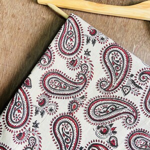 Op de afbeelding: Een witte stof met een paisley-patroon in tinten rood en bruin. De stof is gevouwen en gedrapeerd over een houten hanger.