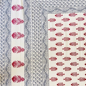 May include: A white textile with a repeating pattern of red and navy blue designs. The design includes floral and leaf motifs, with a border of paisley and geometric shapes. The overall aesthetic is traditional and decorative.