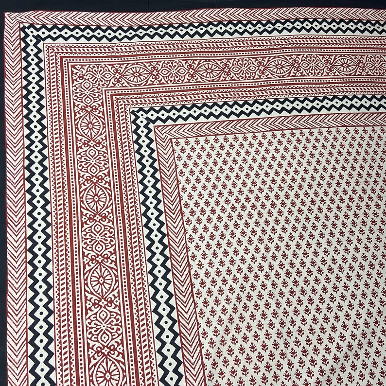 Puede incluir: Una tela de algod&oacute;n blanca con un dise&ntilde;o estampado en bloque rojo y negro. La tela presenta un patr&oacute;n floral repetitivo en el centro y un borde con motivos geom&eacute;tricos y florales.