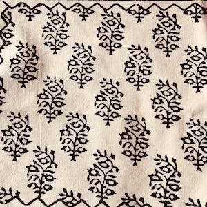 May include: Close-up of a cream-colored fabric with a repeating pattern of dark brown floral motifs. The design includes a border of zigzags and dots in a darker shade of brown. The fabric has a textured appearance.