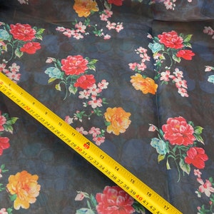 Puede incluir: Un primer plano de una tela azul oscuro transparente con estampado floral. La tela presenta flores rojas, amarillas y blancas sobre un fondo azul oscuro. Una cinta m&eacute;trica amarilla es visible en la esquina inferior derecha de la imagen.